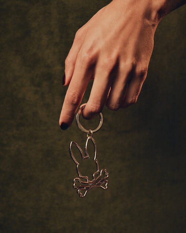 A black-polished hand holds the OUTLINE BUNNY KEYCHAIN - B6A565E200 by Psycho Bunny, featuring a gold alloy bunny skull and crossbones against a dark green background. This intriguing accessory sparks curiosity and conversation.