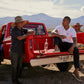 Three men by a red pickup truck in a rural setting with mountains in the background. One man, wearing a straw hat and a Psycho Bunny MENS CLASSIC POLO - B6K001ARPC, is standing and talking, while another sits