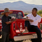 An older man wearing a straw hat and a younger man in a white Psycho Bunny MENS CLASSIC POLO - B6K001ARPC lean on the tailgate of a red Ford pickup truck in a rural landscape, casually chatting with soft voices.