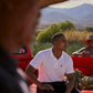 A young man in a Psycho Bunny Pima cotton polo shirt sits on the tailgate of a red pickup truck, listening attentively. Two men wearing straw hats are in the background near a pile of fresh corn.