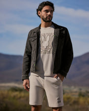 A man stands outdoors in front of a mountainous landscape. He is wearing a black Psycho Bunny MENS PATHE DENIM JACKET - B6J741X1CO with adjustable cuffs over a white t-shirt featuring a faded bunny skull and crossbones design, and matching white shorts. His hands are in his shorts pockets, and he has a thoughtful expression.