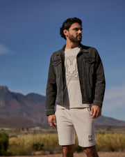A man with a beard is standing outdoors against a backdrop of mountains and a clear blue sky. He is wearing a Psycho Bunny MENS PATHE DENIM JACKET - B6J741X1CO over a white T-shirt featuring a faded bunny design and white shorts with a small bunny logo. The jacket has adjustable cuffs, adding to its rugged appeal.