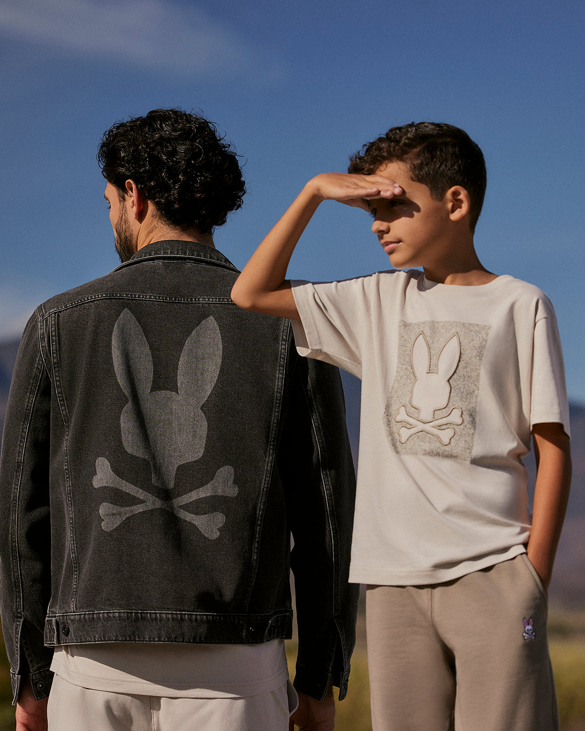 A man and a boy stand outdoors under a clear blue sky with mountains in the background. The man wears a Psycho Bunny MENS PATHE DENIM JACKET - B6J741X1CO with adjustable cuffs, featuring a Bunny logo rabbit skull graphic on the back. The boy wears a matching t-shirt with a similar graphic and shades his eyes with his hand.