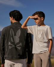 A man and a boy stand outdoors under a clear blue sky with mountains in the background. The man wears a Psycho Bunny MENS PATHE DENIM JACKET - B6J741X1CO with adjustable cuffs, featuring a Bunny logo rabbit skull graphic on the back. The boy wears a matching t-shirt with a similar graphic and shades his eyes with his hand.