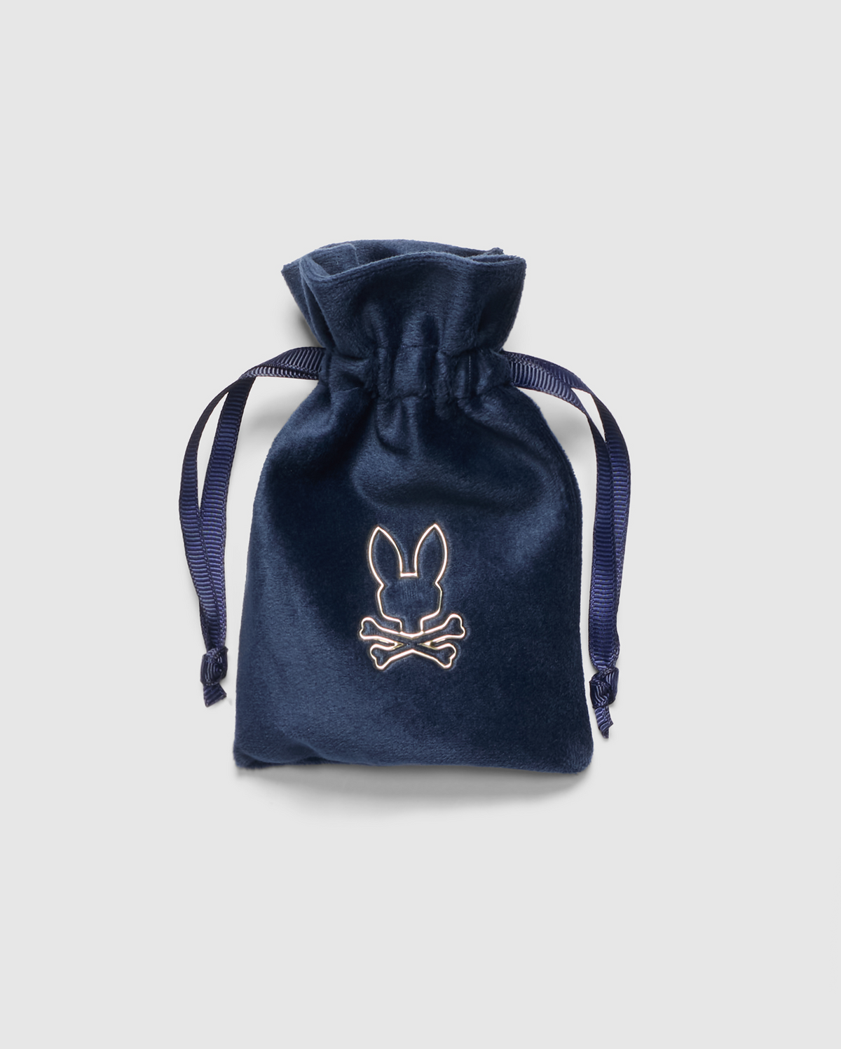 The PB ACCESSORY VELVET BAG - PBVLTBAG by Psycho Bunny is a navy blue velvet drawstring pouch featuring an embroidered rabbit and crossbones logo on the front. It includes dark blue ribbon ties and is showcased against a light gray background.