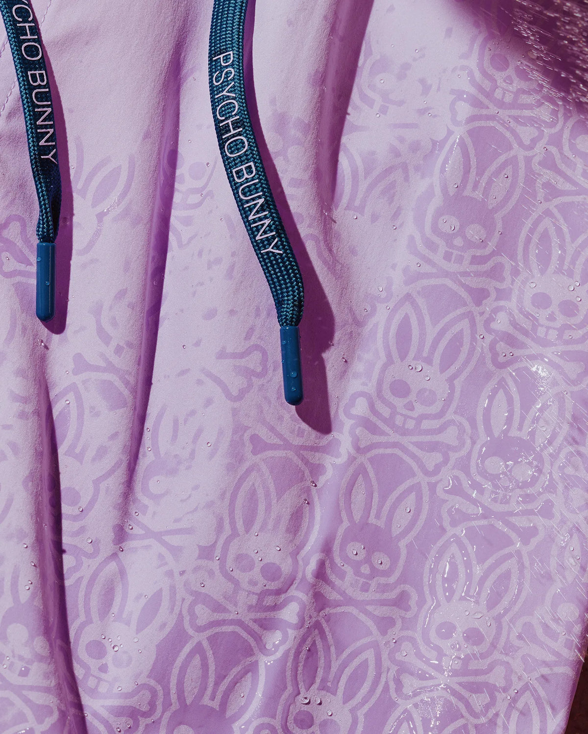 Mens Samos Hydrochromic Printed Swim Trunk in lavender, featuring an all-over subtle bunny skull and crossbones hydrochromic print, visible with water droplets. Dark teal drawstrings with Psycho Bunny text and blue tips.