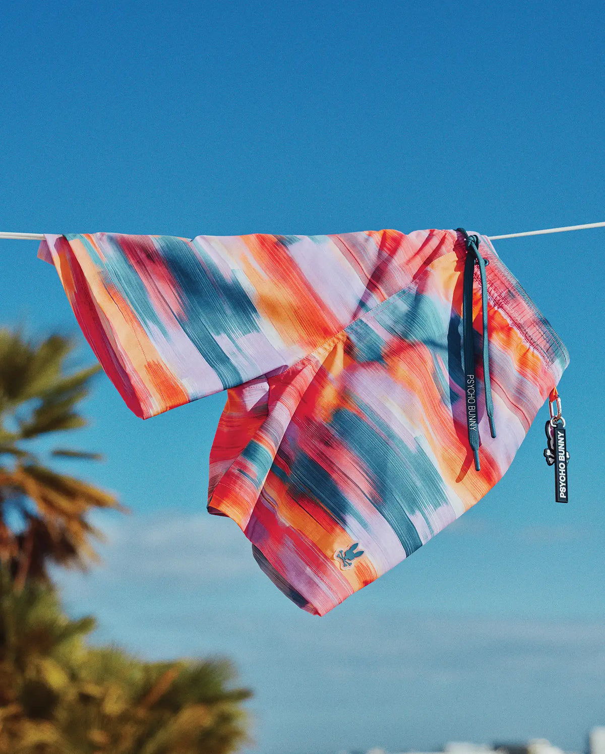 Mens Raphael Swim Trunk with vibrant abstract brushstroke print in orange, pink, blue, purple, and red. Features a relaxed fit, smooth quick-dry fabric, teal drawstring with Psycho Bunny text, and an embroidered bunny skull logo.