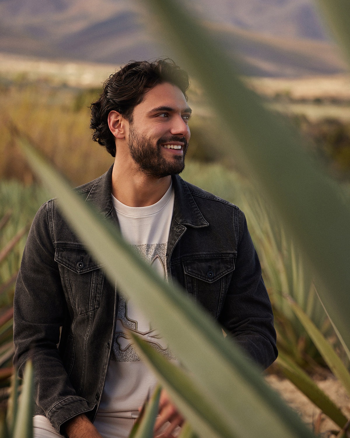 A man with dark hair and a beard smiles while sitting outdoors among tall, green plants. He is wearing a Psycho Bunny MENS PATHE DENIM JACKET - B6J741X1CO with adjustable cuffs over a white t-shirt. The background features a hilly landscape with mountains in the distance.