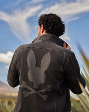 A person with curly hair faces away from the camera, adjusting the collar of a denim jacket. The jacket, with adjustable cuffs, features a large design on the back depicting a stylized bunny head with crossed bones underneath. The background shows a clear sky with wispy clouds. The Psycho Bunny MENS PATHE DENIM JACKET - B6J741X1CO stands out prominently in this serene setting.
