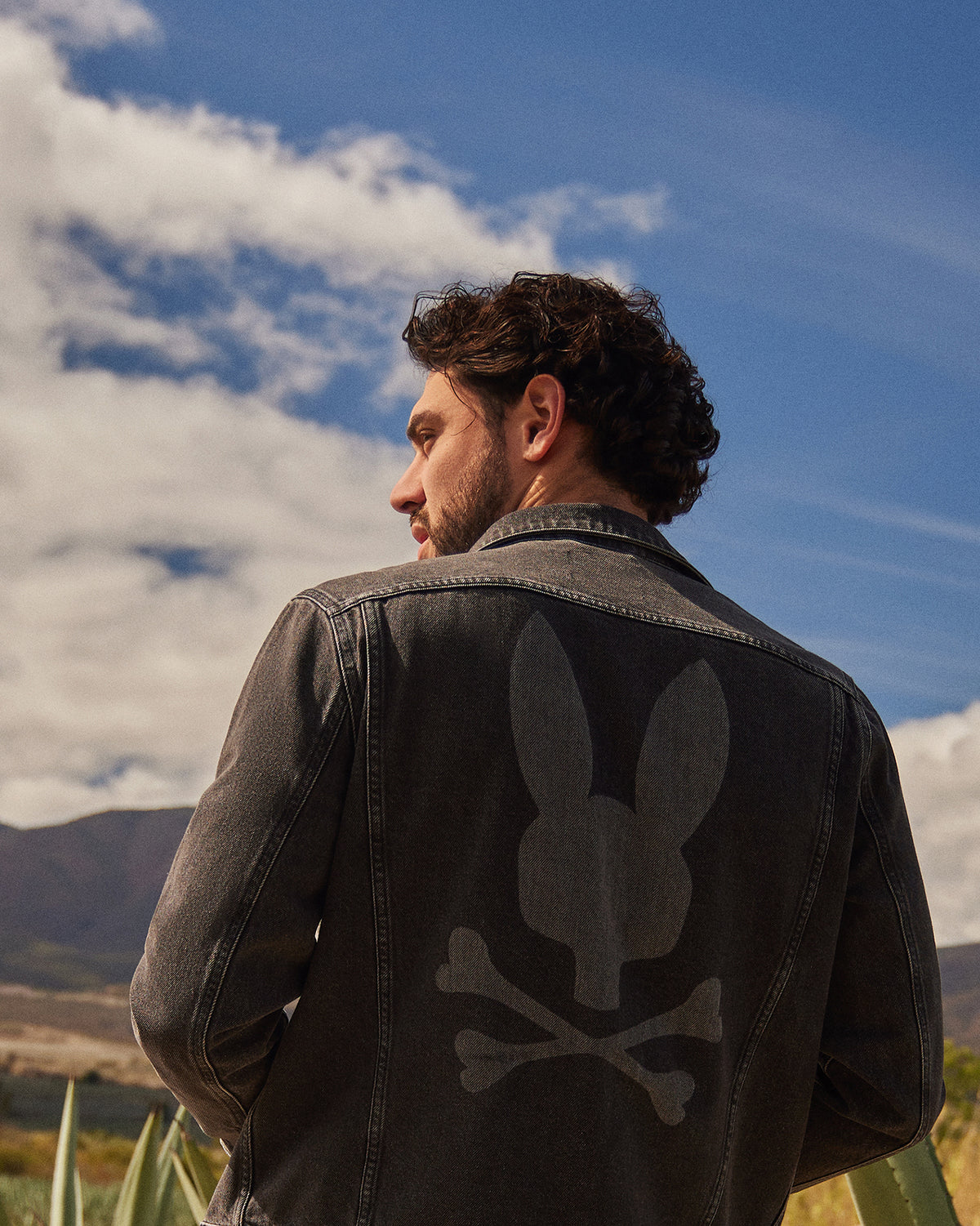 A man with curly hair stands outdoors, looking to his right, wearing a Psycho Bunny MENS PATHE DENIM JACKET - B6J741X1CO with adjustable cuffs and a large faded bunny head and crossbones design on the back. The sky is bright with scattered clouds, and a mountain range is visible in the background.