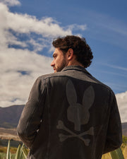 A man with curly hair stands outdoors, looking to his right, wearing a Psycho Bunny MENS PATHE DENIM JACKET - B6J741X1CO with adjustable cuffs and a large faded bunny head and crossbones design on the back. The sky is bright with scattered clouds, and a mountain range is visible in the background.