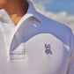 Close-up of a person wearing a white Psycho Bunny Pima cotton polo shirt with a small bunny logo embroidered on the chest. The background shows a clear blue sky with fluffy white clouds.