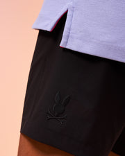 Close-up of a person wearing Psycho Bunny MENS WILLIS STRETCH TENCEL SHORT - B6R239Y1WB with an embroidered skull and bunny ears logo near the hem, and a light purple shirt with slits at the sides. The background is a soft peach color, creating a striking contrast that highlights the comfortable men's trousers in this stylish ensemble.