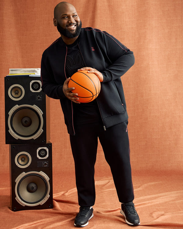 A bearded man wearing Psycho Bunny's Big and Tall Travis Track Pants holds a basketball, smiling and relaxed beside large speakers against a brownish-orange background.