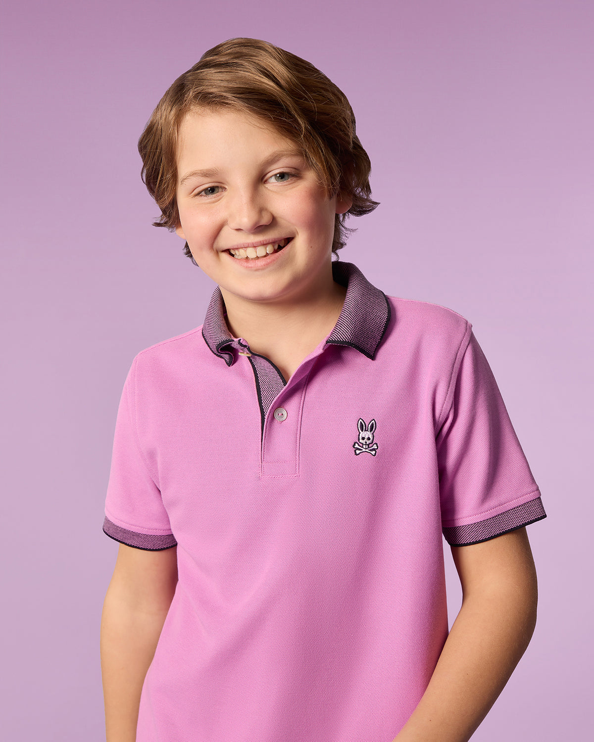 A young boy with light brown hair smiles at the camera. He is wearing a pink Psycho Bunny KIDS SOUTHPORT PIQUE POLO SHIRT - B0K263B200 made of soft Pima cotton, featuring grey trim and an embroidered bunny logo on the front. The background is a soft purple color.