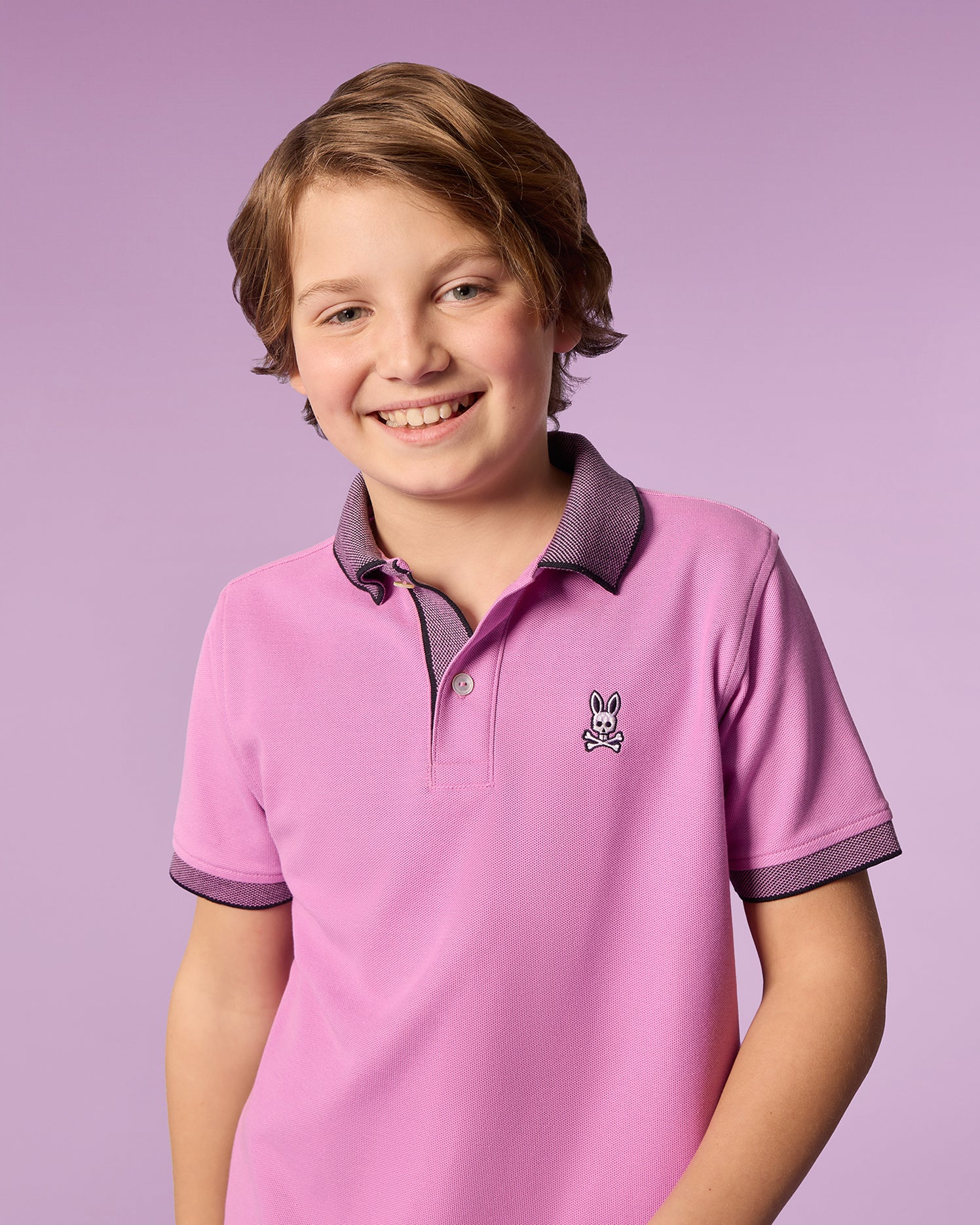 A young boy with light brown hair smiles at the camera. He is wearing a pink Psycho Bunny KIDS SOUTHPORT PIQUE POLO SHIRT - B0K263B200 made of soft Pima cotton, featuring grey trim and an embroidered bunny logo on the front. The background is a soft purple color.