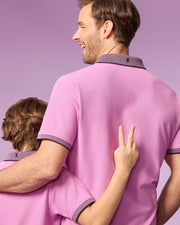 A man and a child stand with their backs to the camera, both wearing matching pink Psycho Bunny KIDS SOUTHPORT PIQUE POLO SHIRT - B0K263B200 with dark collars. The man has his arm around the child's shoulders, while the child makes a peace sign with their hand. The background is a solid, light purple color.