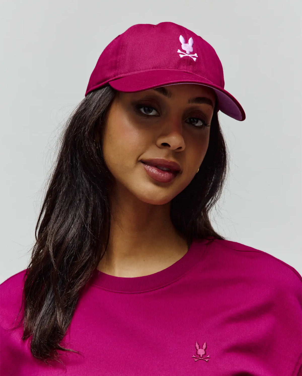 Womens Amaris Baseball Cap in magenta with an embroidered light pink Psycho Bunny skull logo, and a matching magenta crewneck top featuring a small embroidered Psycho Bunny logo on the left chest.