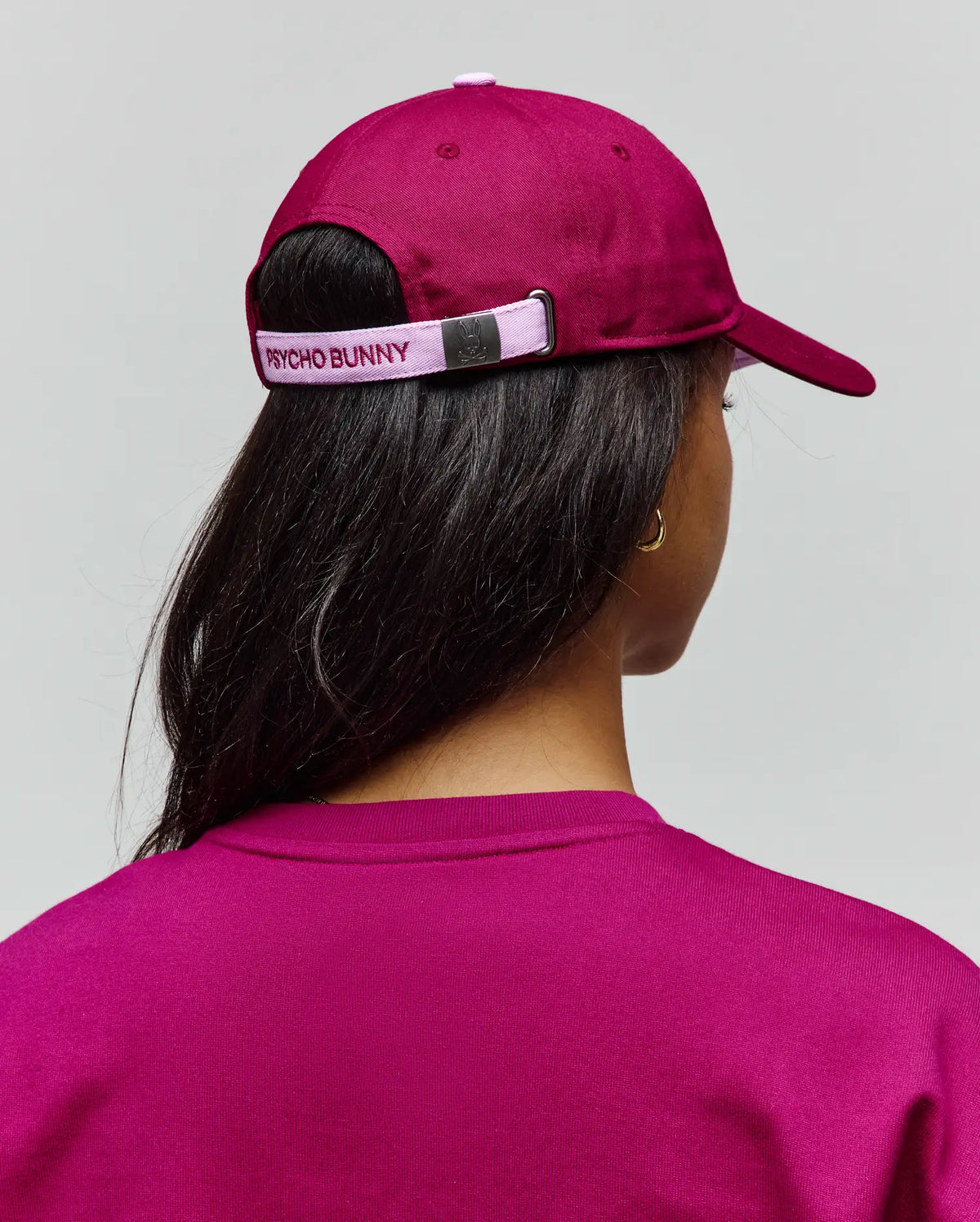 Womens Amaris Baseball Cap in deep magenta cotton twill with a light pink adjustable strap featuring embroidered PSYCHO BUNNY text and a metallic bunny skull buckle, paired with a matching magenta knit crewneck top.
