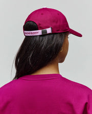 Womens Amaris Baseball Cap in deep magenta cotton twill with a light pink adjustable strap featuring embroidered PSYCHO BUNNY text and a metallic bunny skull buckle, paired with a matching magenta knit crewneck top.