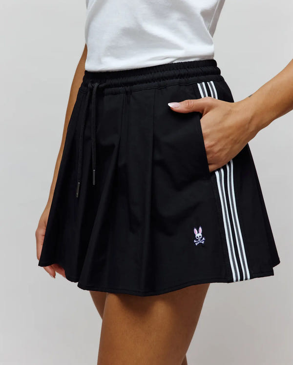 Womens Cecilia Sport Skort in black, featuring a relaxed fit, smooth fabric, pleated design, elastic drawstring waist, side pockets, three white stripes on the right, and a small embroidered pink and white Psycho Bunny skull logo on the lower left front.