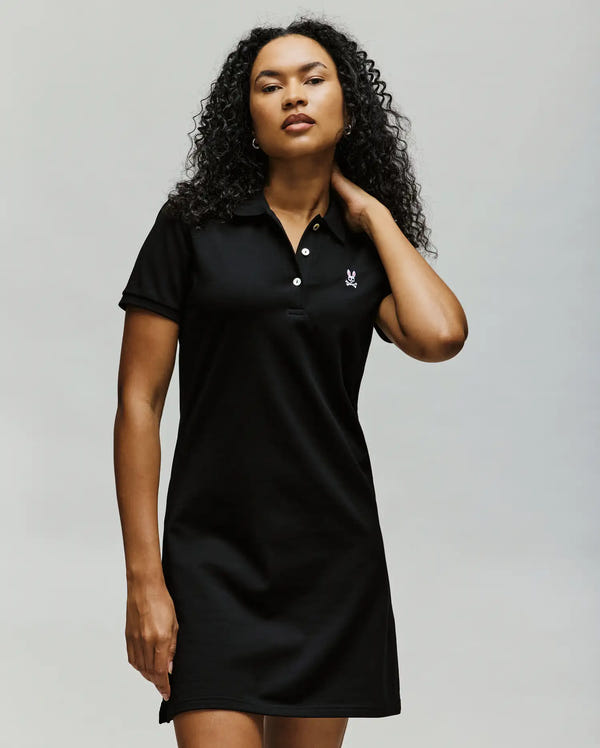 A woman with curly hair wears the Psycho Bunny WOMENS POLO DRESS - WO2000003, featuring a short sleeve design and embroidered Bunny logo on the chest, standing against a plain light background and touching her neck.