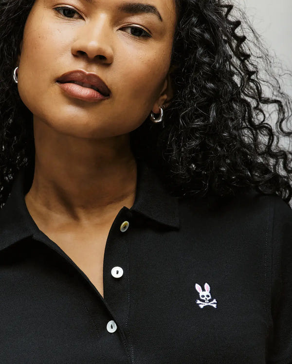 A person with curly hair wears silver hoop earrings and a black Psycho Bunny WOMENS POLO DRESS (WO2000003) featuring a small pink and white bunny logo with crossbones on the chest.