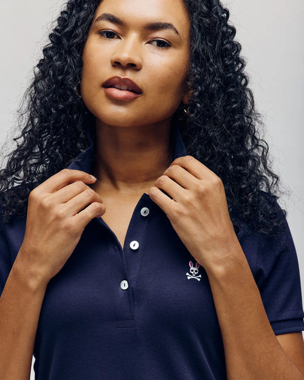 A woman with curly dark hair wears the Psycho Bunny WOMENS POLO DRESS - WO2000003 in navy blue, featuring white buttons, a small skull and crossbones logo, and a feminine silhouette, made from 100% Pima cotton. She holds the collar against a neutral background.