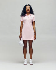 A woman with curly hair wears the Psycho Bunny WOMENS POLO DRESS - WO2000003 in light pink with Mother-of-Pearl buttons and white sneakers, holding a white cap against a plain, light background.
