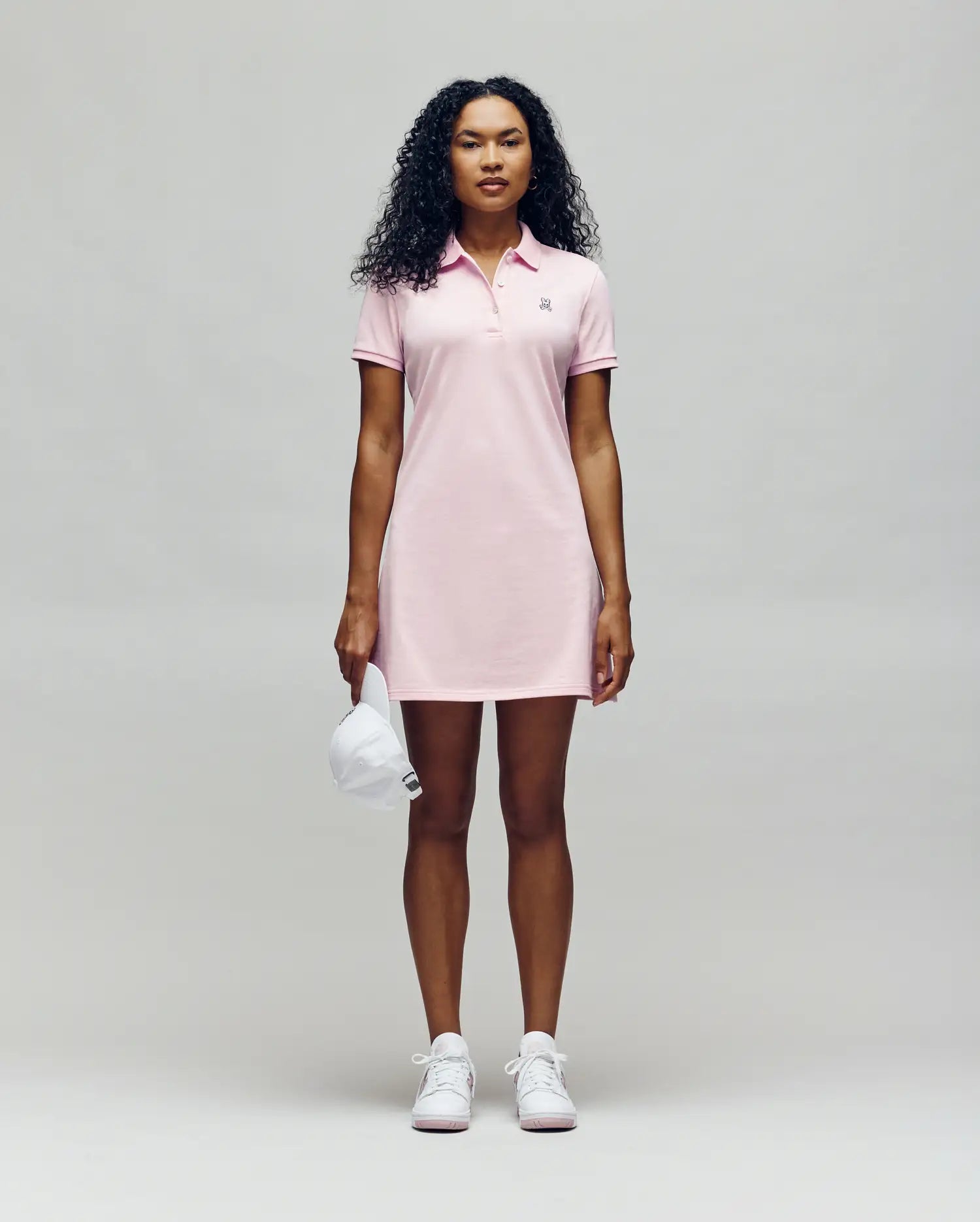 A woman with curly hair wears the Psycho Bunny WOMENS POLO DRESS - WO2000003 in light pink with Mother-of-Pearl buttons and white sneakers, holding a white cap against a plain, light background.