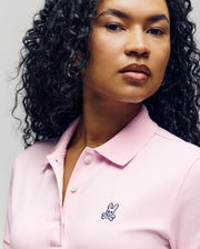 A woman with long curly hair wears a light pink Psycho Bunny WOMENS POLO DRESS (WO2000003) featuring an embroidered skull-and-crossbones bunny logo and Mother-of-Pearl buttons, set against a plain, neutral background.