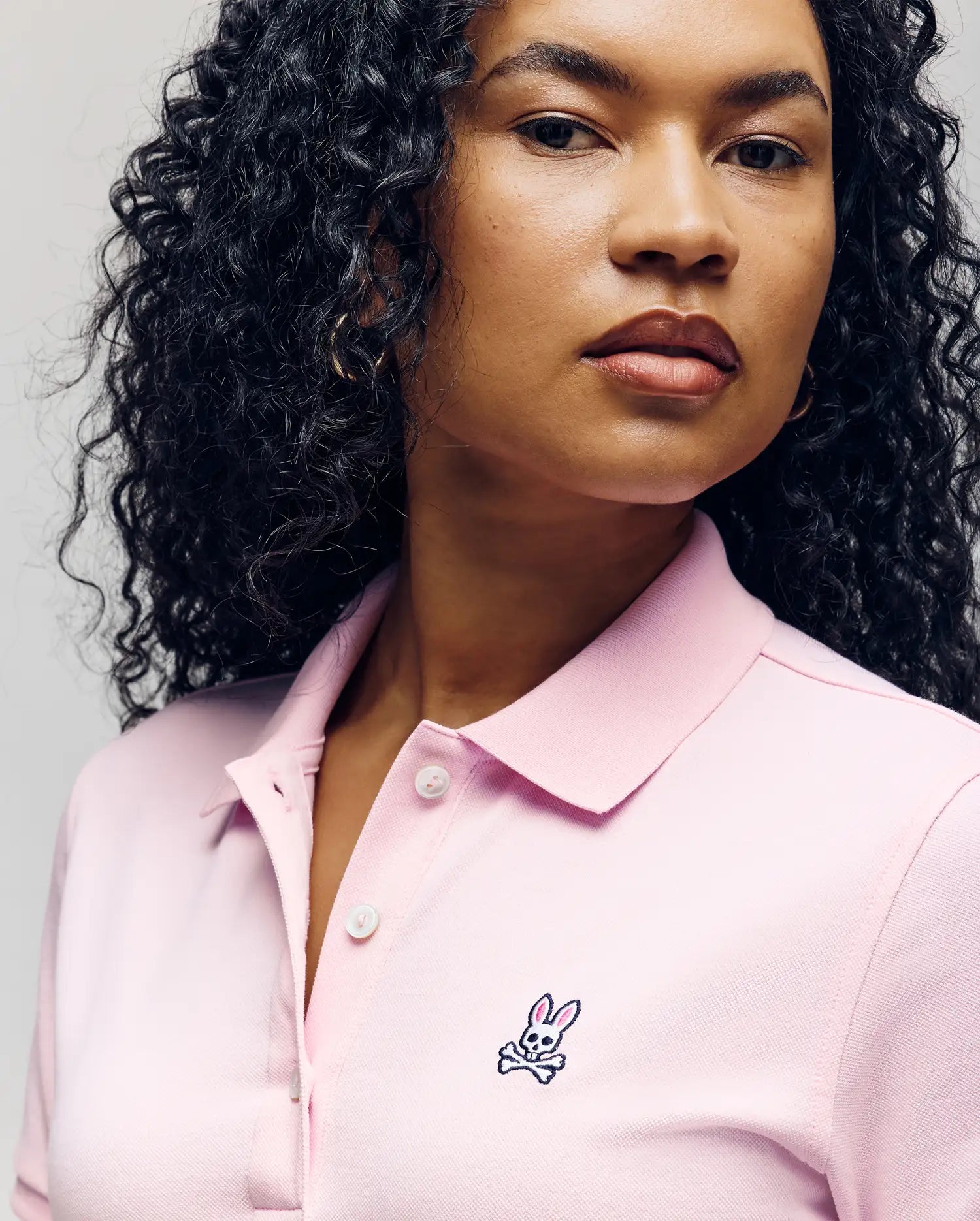 A woman with long curly hair wears a light pink Psycho Bunny WOMENS POLO DRESS (WO2000003) featuring an embroidered skull-and-crossbones bunny logo and Mother-of-Pearl buttons, set against a plain, neutral background.