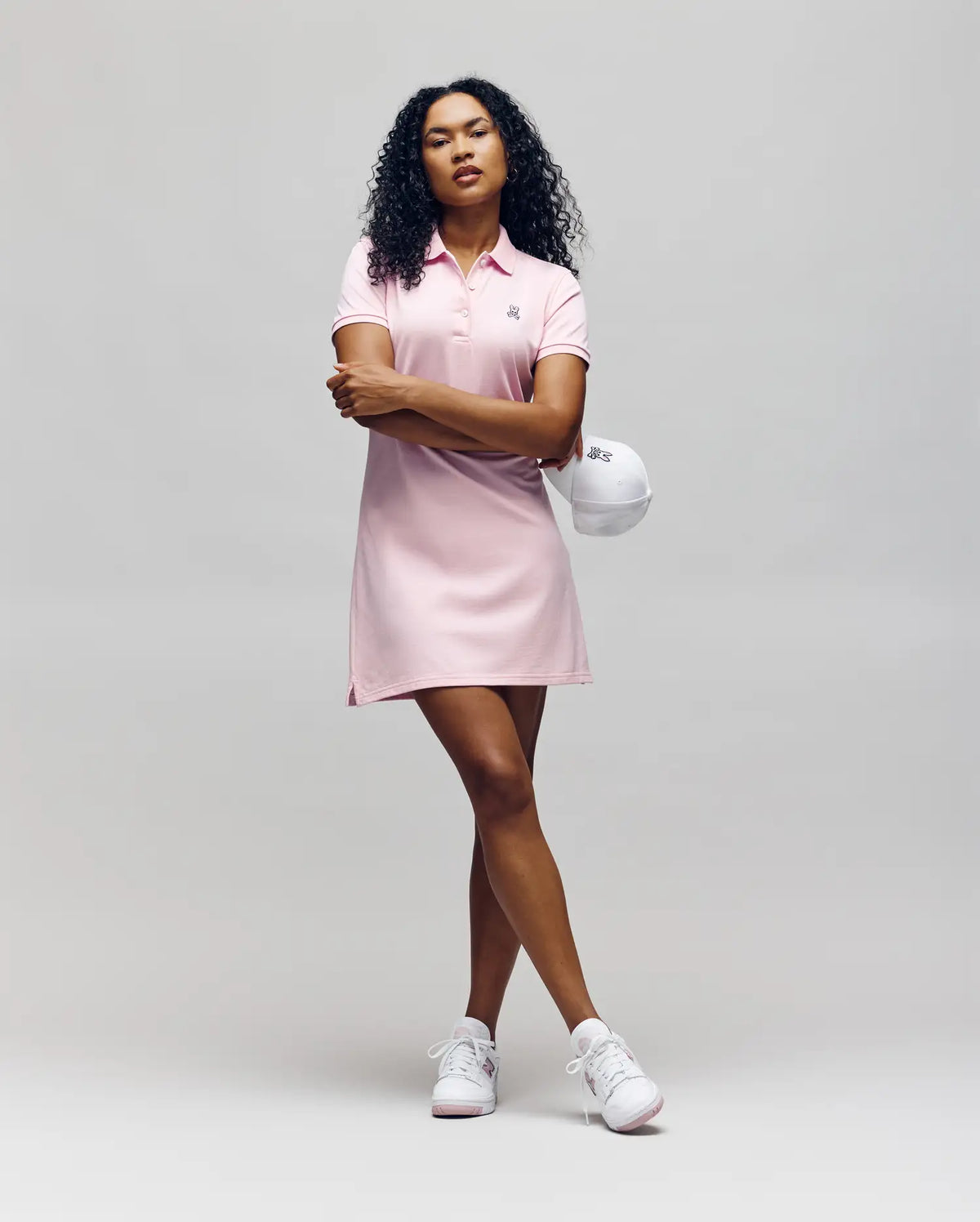 A person with curly hair wears the Psycho Bunny WOMENS POLO DRESS - WO2000003 in light pink, styled with white sneakers and a white cap, standing with arms crossed against a plain, light gray background.