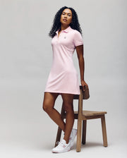 A person with curly hair poses confidently beside a wooden chair, wearing Psycho Bunny’s WOMENS POLO DRESS - WO2000003 in light pink with Mother-of-Pearl buttons and white sneakers, against a plain background.
