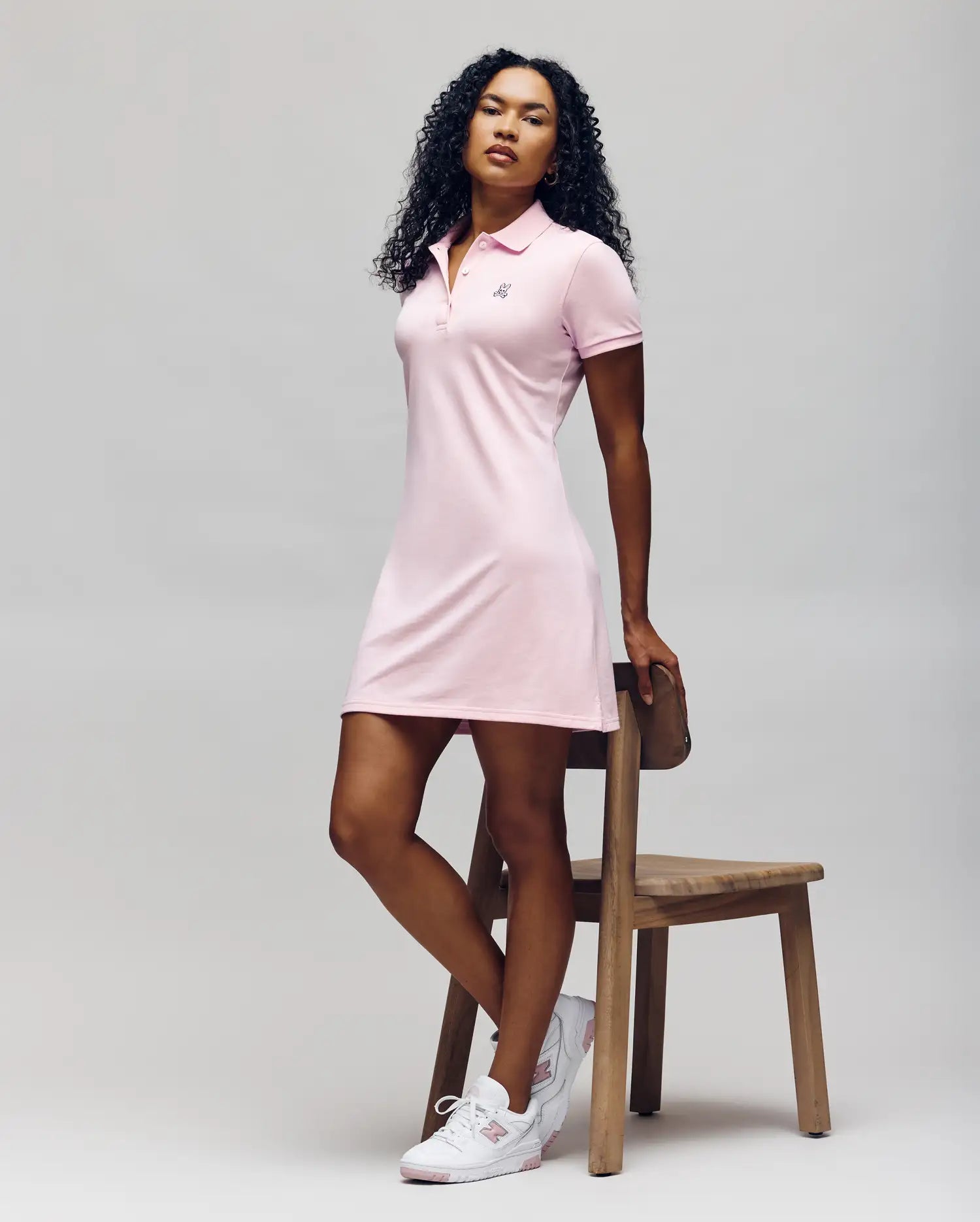 A person with curly hair poses confidently beside a wooden chair, wearing Psycho Bunny’s WOMENS POLO DRESS - WO2000003 in light pink with Mother-of-Pearl buttons and white sneakers, against a plain background.