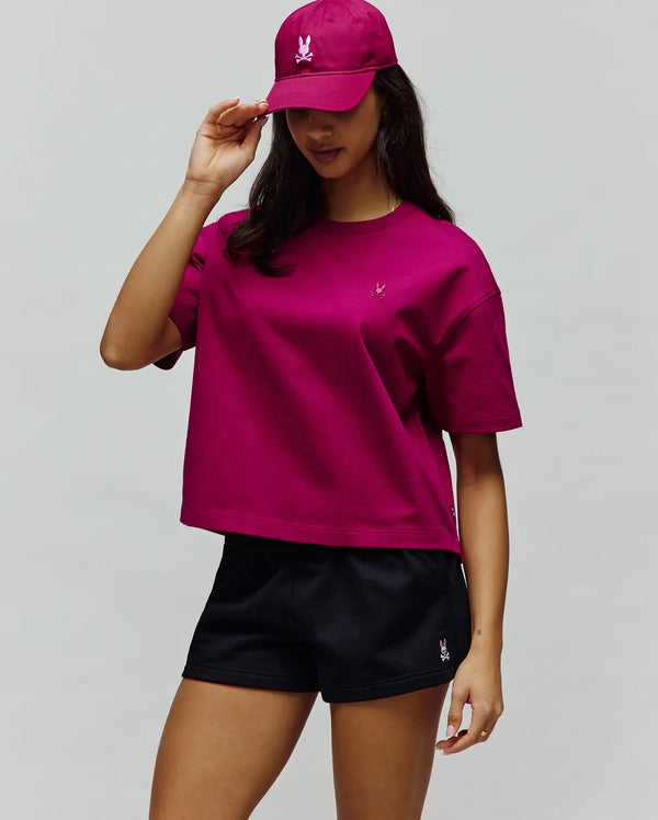 Magenta Women's Camille Oversized Fit Tee, a relaxed jersey top with a small pink embroidered Psycho Bunny chest logo. Black athletic shorts and a matching magenta twill cap also feature embroidered Psycho Bunny logos.