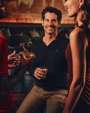 A man in a dark polo smiles while holding a drink in a lively bar setting, surrounded by two people enjoying their beverages and conversation. The warmth of the room perfectly complements his Psycho Bunny Men's Johnny Collar Sweater (B6E258E200), blending seamlessly with the blurred seating and decor behind them.