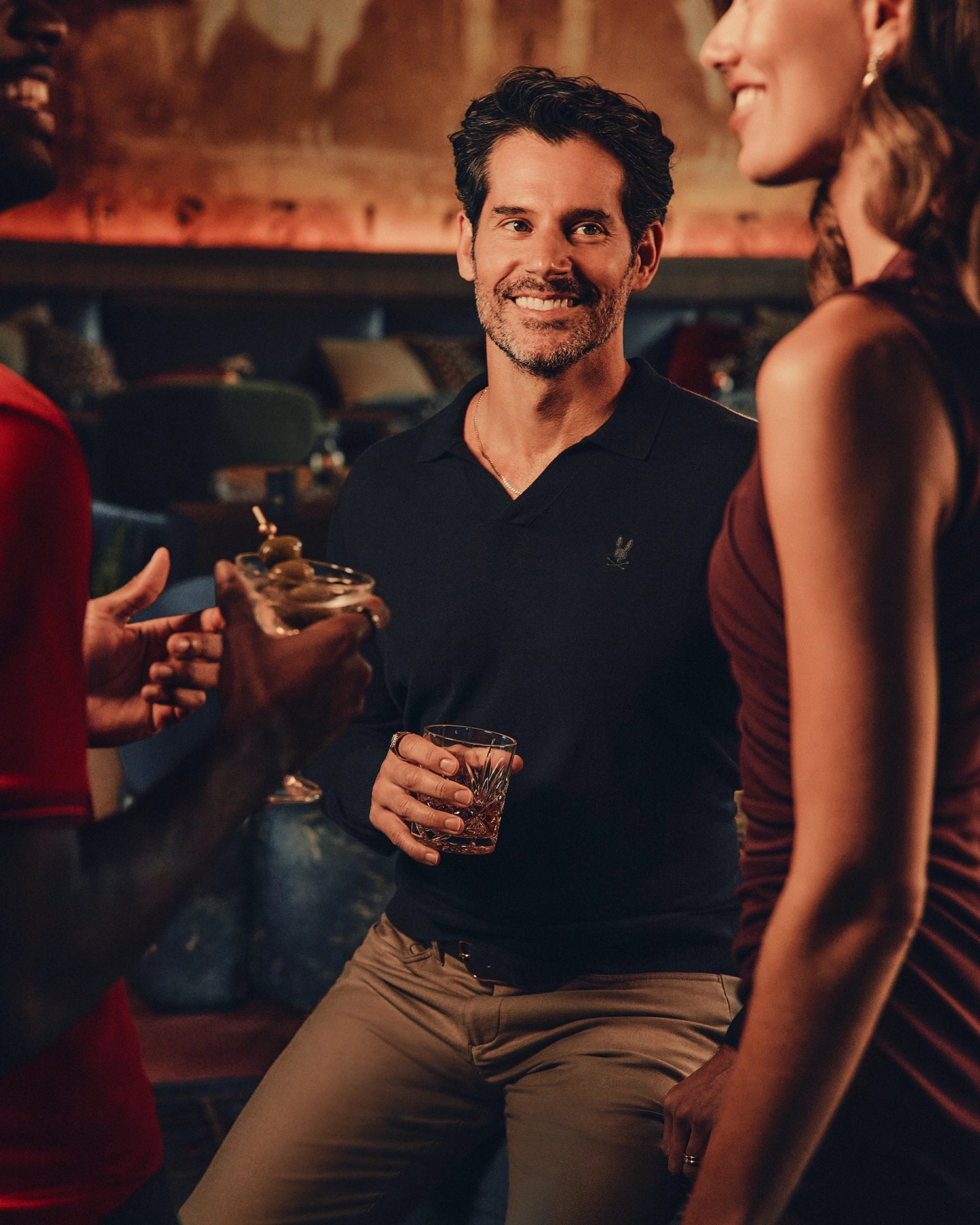 A man in a dark polo smiles while holding a drink in a lively bar setting, surrounded by two people enjoying their beverages and conversation. The warmth of the room perfectly complements his Psycho Bunny Men's Johnny Collar Sweater (B6E258E200), blending seamlessly with the blurred seating and decor behind them.