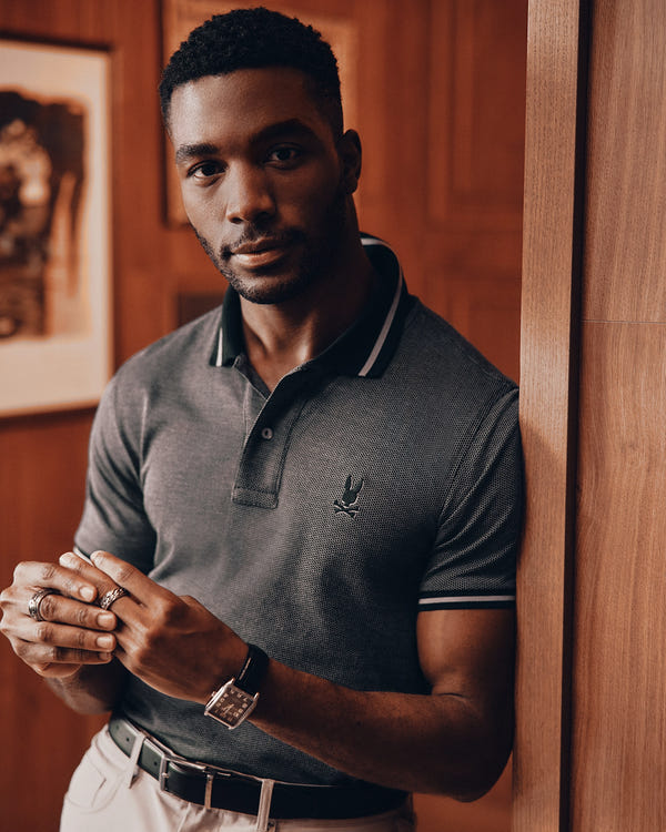 A man with short hair and a beard is leaning against a wooden wall, dressed in a dark Psycho Bunny MENS WARSAW JACQUARD PIQUE POLO - B6K901E200 made from Peruvian Pima cotton, light pants, and a watch. He has rings on his fingers and gazes directly at the camera, with framed pictures in the background.