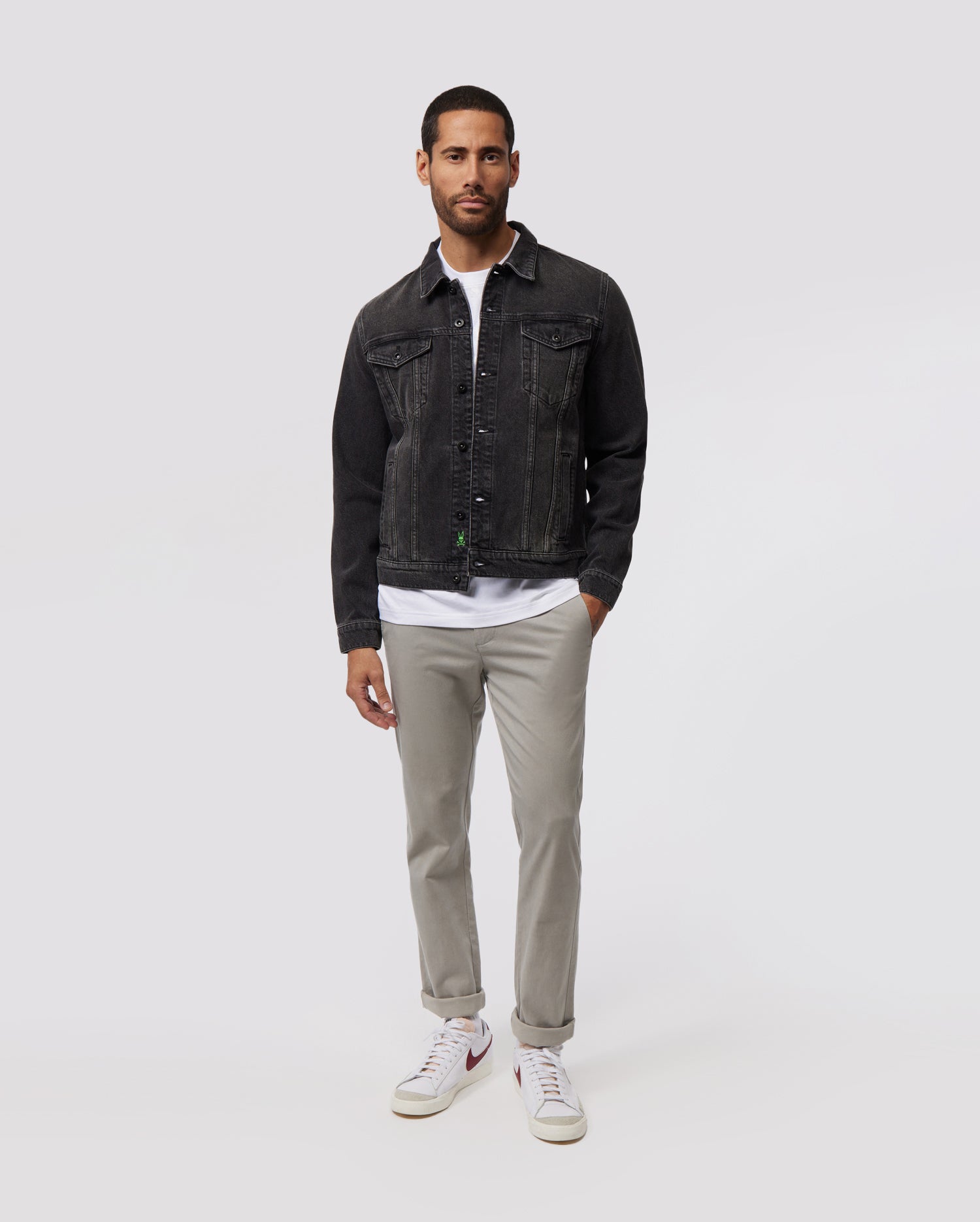 A man wearing a dark denim Psycho Bunny MENS PATHE DENIM JACKET - B6J741X1CO with adjustable cuffs over a white shirt, light grey pants, and white sneakers stands against a plain, light grey background.