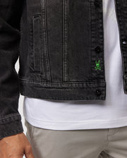 A close-up of a person's lower torso shows them wearing a Psycho Bunny MENS PATHE DENIM JACKET - B6J741X1CO over a white shirt and light gray pants. The jacket, featuring adjustable cuffs, has a small green embroidered bunny near the front buttons. The person's left hand is partially visible along the left edge.