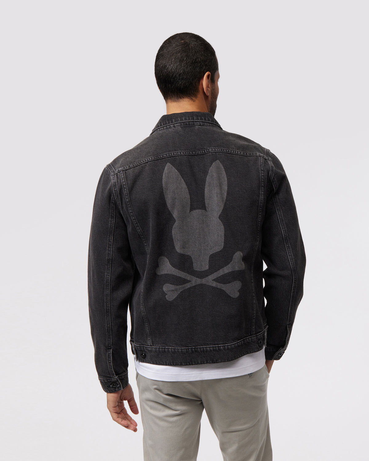 A man with short dark hair is seen from the back, wearing a MENS PATHE DENIM JACKET - B6J741X1CO by Psycho Bunny featuring a large gray Bunny logo of a bunny skull and crossbones on the back. The jacket also has adjustable cuffs. He is also wearing light gray pants and a white shirt underneath. The background is plain light gray.
