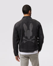 A man with short dark hair is seen from the back, wearing a MENS PATHE DENIM JACKET - B6J741X1CO by Psycho Bunny featuring a large gray Bunny logo of a bunny skull and crossbones on the back. The jacket also has adjustable cuffs. He is also wearing light gray pants and a white shirt underneath. The background is plain light gray.