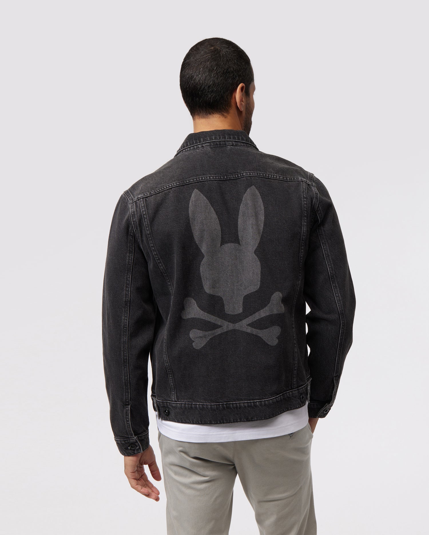 A man with short dark hair is seen from the back, wearing a MENS PATHE DENIM JACKET - B6J741X1CO by Psycho Bunny featuring a large gray Bunny logo of a bunny skull and crossbones on the back. The jacket also has adjustable cuffs. He is also wearing light gray pants and a white shirt underneath. The background is plain light gray.
