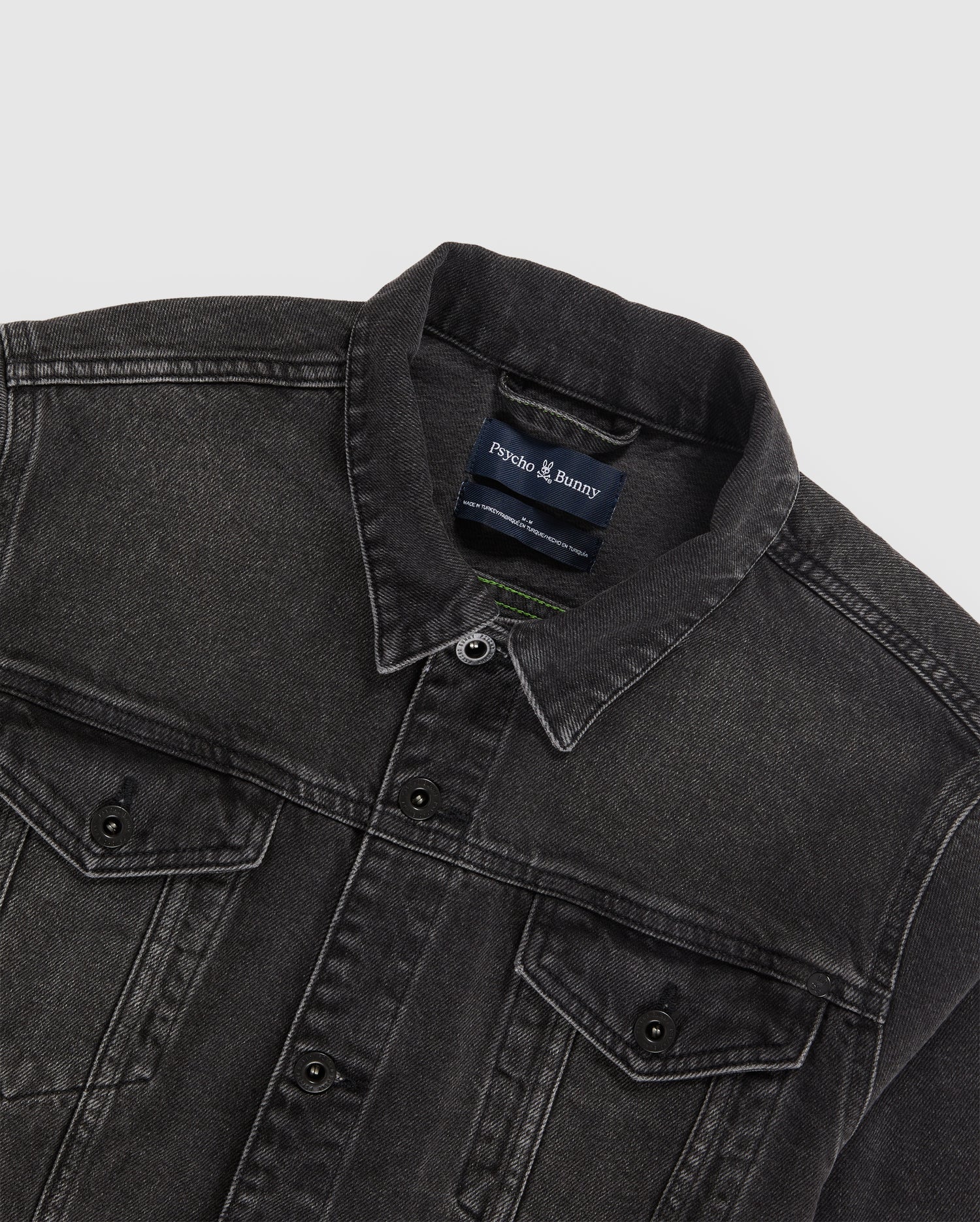 A black MENS PATHE DENIM JACKET - B6J741X1CO with a collar and button-down front is spread out flat. The jacket features two chest pockets with buttoned flaps, adjustable cuffs, and visible stitching. The brand tag inside the collar reads "Psycho Bunny." The background is plain white.
