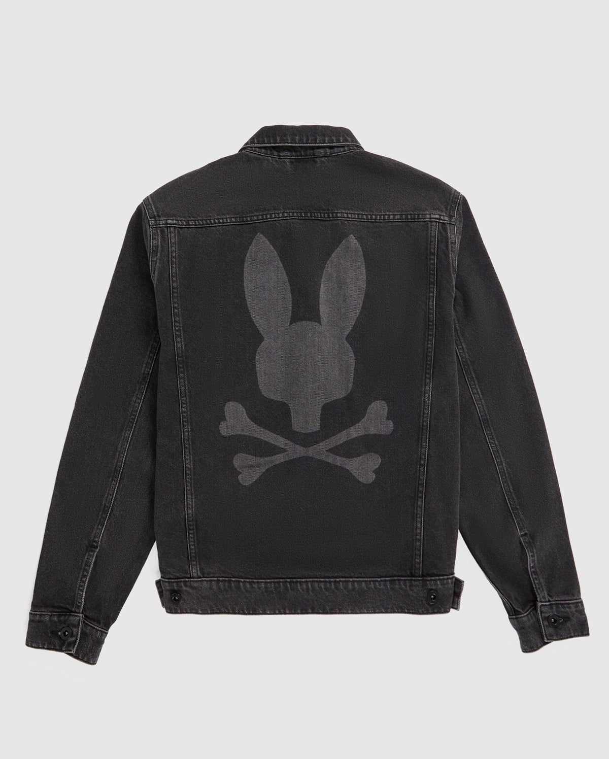 A black Psycho Bunny MENS PATHE DENIM JACKET - B6J741X1CO with a large, faded bunny graphic on the back depicting a bunny head above two crossed bones. The jacket features a button-up front, adjustable cuffs, and visible stitching accentuating the seams and edges. The background is plain white.