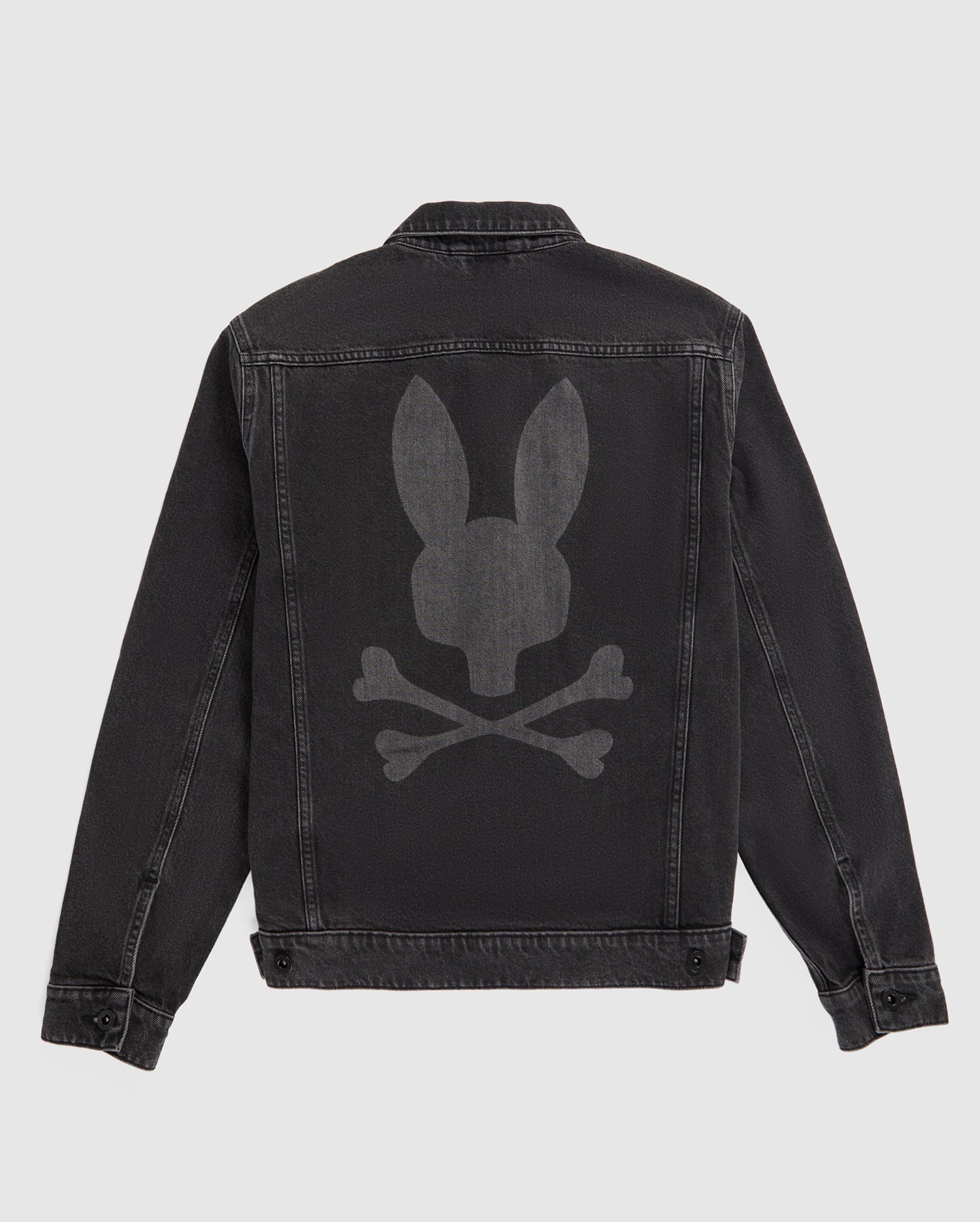 A black Psycho Bunny MENS PATHE DENIM JACKET - B6J741X1CO with a large, faded bunny graphic on the back depicting a bunny head above two crossed bones. The jacket features a button-up front, adjustable cuffs, and visible stitching accentuating the seams and edges. The background is plain white.