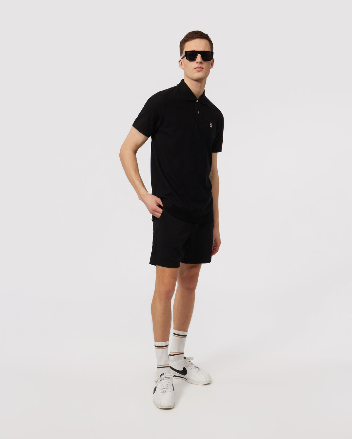 A man wearing a black polo shirt, Psycho Bunny eco-friendly shorts, white socks, and white sneakers, accessorized with sunglasses, stands posing against a plain white background.