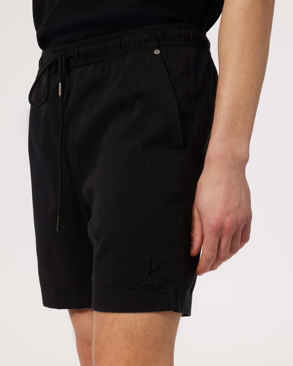 Close-up of a person wearing black Psycho Bunny eco-friendly shorts with a drawstring, featuring a small white logo on the left leg.