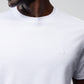 Close-up of a person wearing a white MENS OUTLINE TEE - B6U500ARPC from the Psycho Bunny collection, featuring an embroidered emblem near the chest area. The emblem showcases a bunny above two crossed bones. The person's head is slightly turned, and the background is a plain light gray.